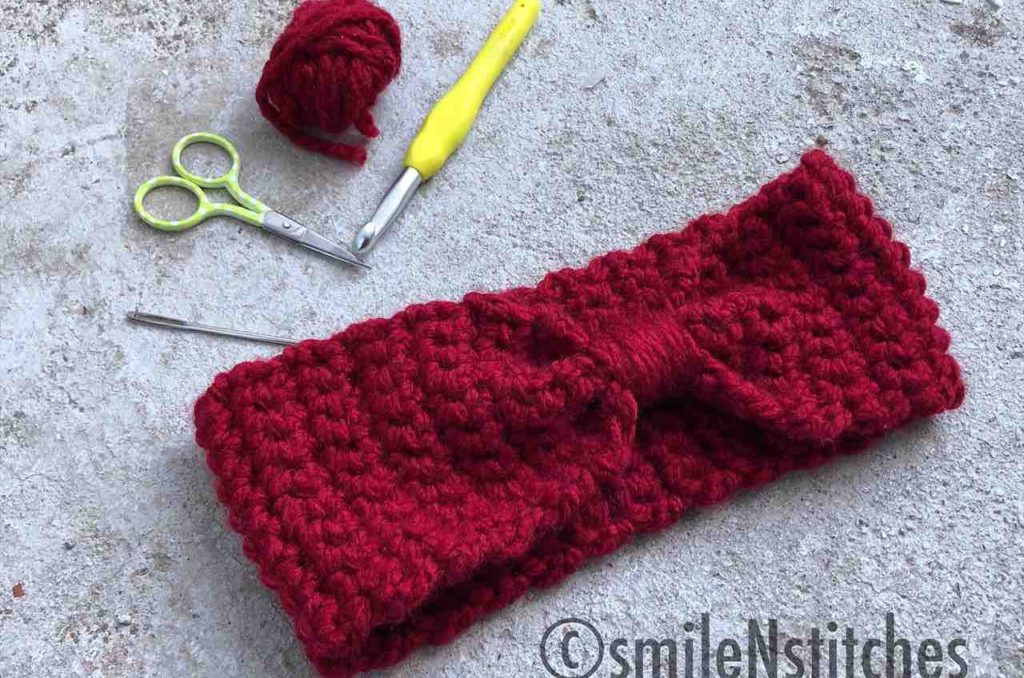 Burgundy bowtie head warmer on a marble background surrounded by crochet tools.