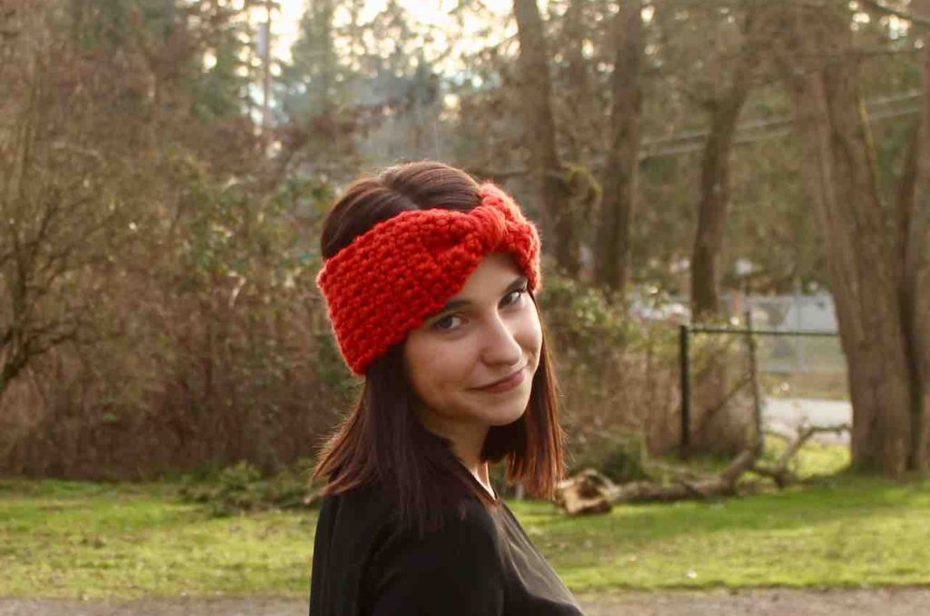 Lovely young lady dressed in black wearing a deep orange chunky head warmer in the midst of luscious  trees and greenery.