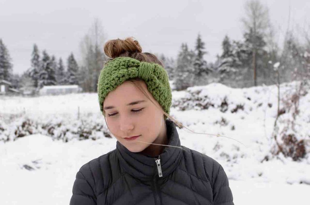 Beautiful blonde young lady with her hair in a bun wearing an apple green chunky bowtie ear warmer in the midst of a snowy field and snow dusted trees.